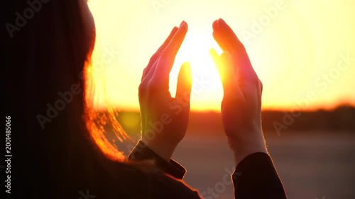 Silhouette of woman raising hands towards glowing sunset, warm sunlight shining through fingers. Hope, mindfulness and inner peace, emotional connection with nature and light calm outdoor moment. Girl