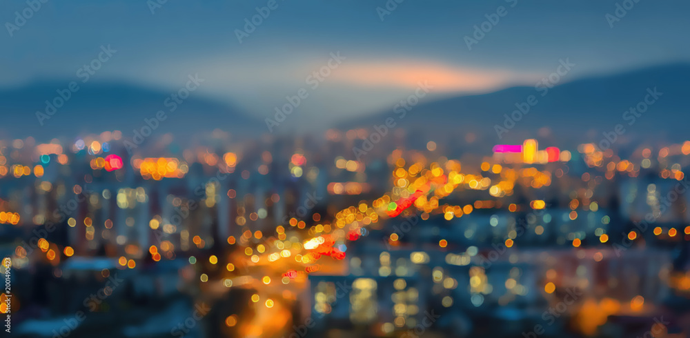 Fototapeta premium Blurred cityscape at dusk with illuminated buildings and distant mountains