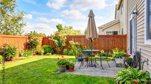 Vászonkép Neat backyard patio with black metal dining set, umbrella, surrounded by green lawn and blooming flowers