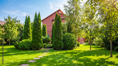 Bild auf Leinwand Green lawn and lush garden with cypress trees and a red brick house under a blue sky on a sunny day