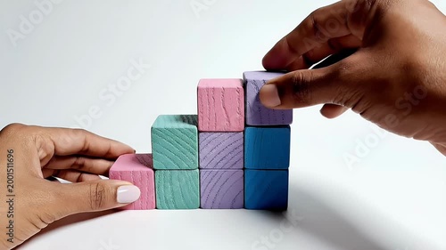 A close-up shot depicts hands carefully stacking colorful wooden blocks, creating a playful and abstract tower against a clean white background.