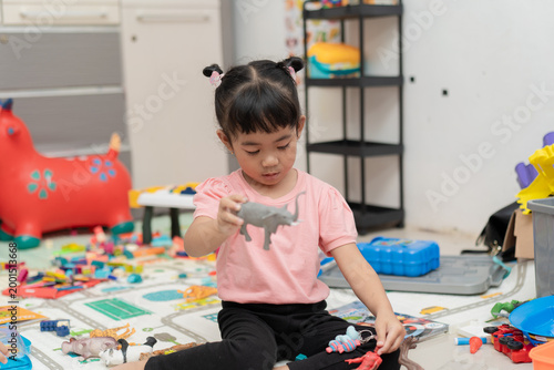 Little Asian girl sitting among many toys holding a grey elephant figurine indoors