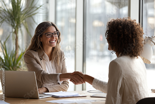 Smiling professional women shaking hands across a modern office desk during a successful job interview and partnership meeting with laptop
