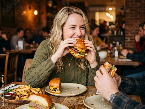Young woman enjoying a large sesame seed burger at a cozy restaurant with friends. Happy blonde girl eating fast food and laughing during a casual dinner date.
