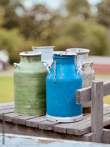 A group of vintage metal milk cans in different colors placed on a rustic wooden table outdoors. The soft blurred background and natural light create a nostalgic rural atmosphere, evoking traditional 