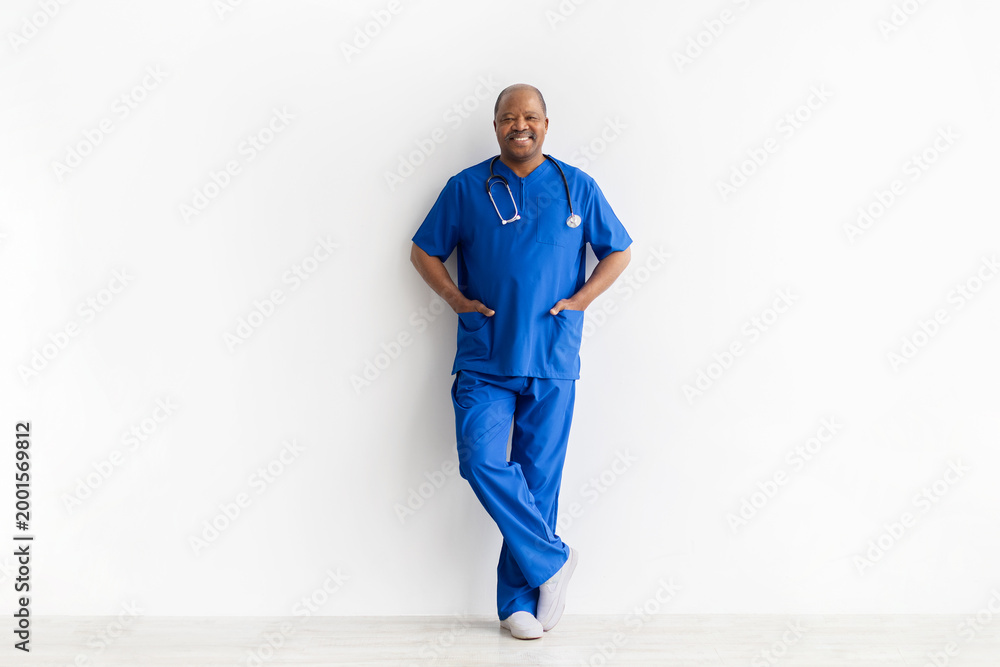 Obraz premium Confident mature African American male doctor in blue scrubs and stethoscope leaning casually against white wall. Full body professional medical portrait on studio background