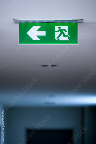 green emergency fire exit sign with arrow and running man symbol hanging on a white ceiling in a dimly lit office building corridor or hallway