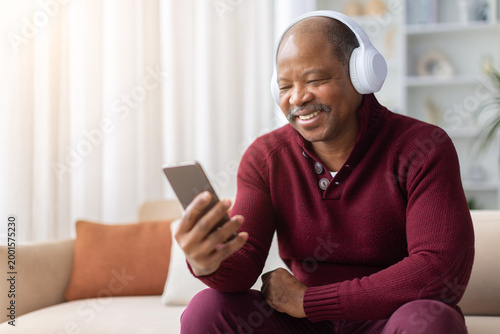 Happy mature African American man wearing wireless headphones, laughing while using smartphone at home. Technology and entertainment lifestyle concept indoors