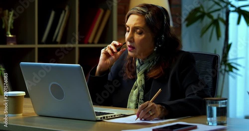Senior Indian businesswoman in corporate attire taking notes while attending training session on laptop in modern cozy office space experienced female executive using headphones with mic at desk