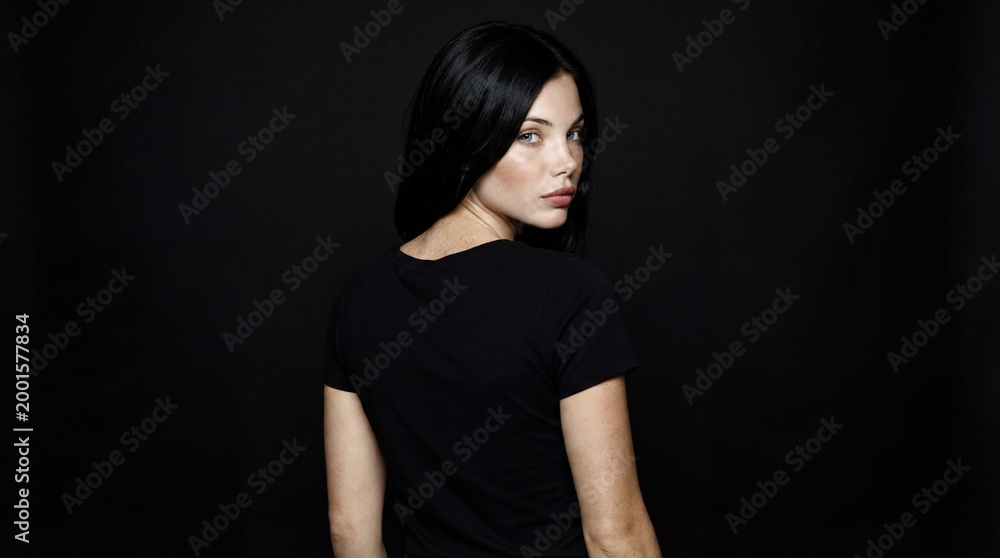 Fototapeta premium A young Caucasian woman with long black hair looks over her shoulder against a dark background. She wears a simple black t-shirt and has a serious expression.
