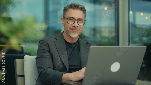 Mature professional man in business casual smiling, working on laptop in contemporary office. Portrait of happy mid adult man with approachable friendly smile. Successful senior entrepreneur.