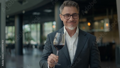 Businessman in suit sitting in a modern restaurant, drinking, tasting red wine. Elegant man holding glass, focusing on evaluating, rating wine. Moment of pleasure and well deserved relaxing break.