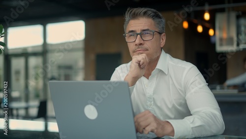 Businessman sitting in a cafe, working on laptop thinking on next business move. Portrait of smart confident mid adut, middle aged man in glasses. Entrepreneur managing business remotely.