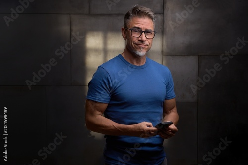 Confident mature man standing against dark industrial wall in office, using mobile phone. Portrait of mid adult handsome male in glasses, smiling. Staying fit and sporty in older age.