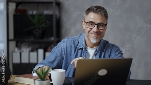 Portrait of mature man with grey hair and beard smiling. Confident businessman working on laptop from home office. Male entrepreneur sitting at desk in bright room wearing casual denim shirt.