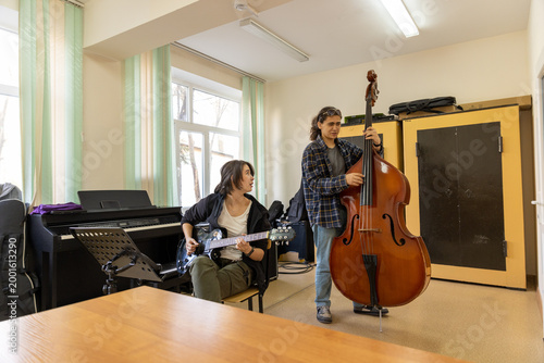 Music college students practicing electric guitar and double bass in a classroom equipped with a piano.