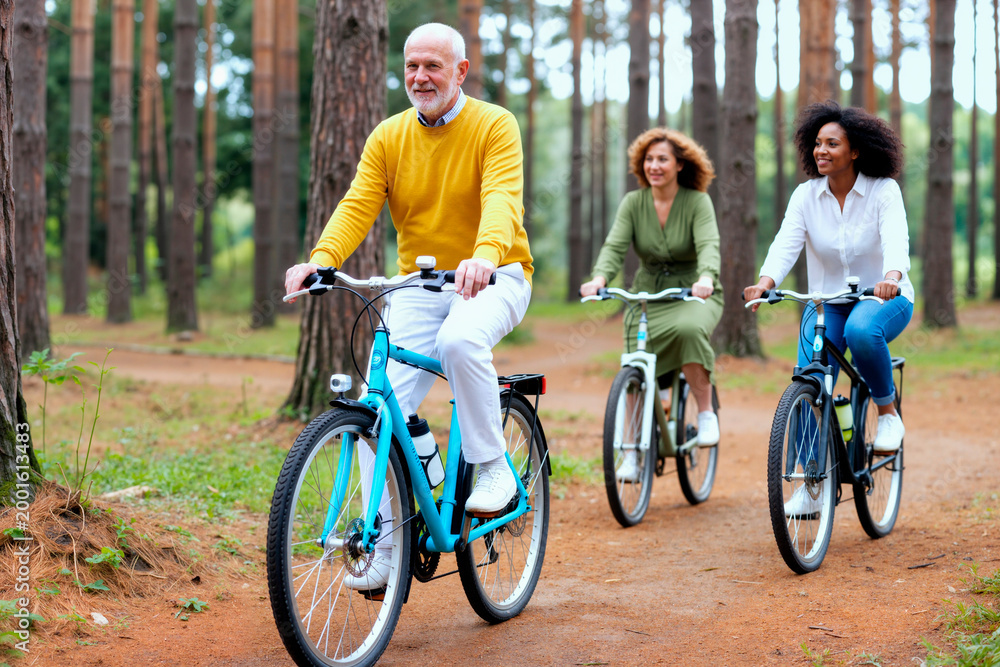 Fototapeta premium Senior Caucasian man leading diverse women riding bicycles on forest trail, promoting active lifestyle and social connection. Ideal for wellness, retirement, recreation marketing