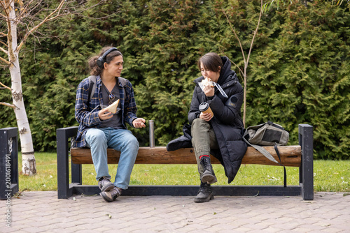 Two students sitting on a wooden bench in a park, eating sandwiches and talking during a break. Student life and outdoor lunch concept.