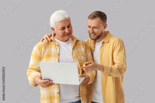 Senior man and his son with laptop on grey background