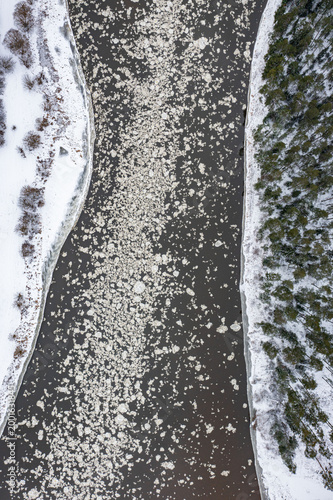 Aerial view of a winter river with floating ice, snowy banks, and forest shoreline