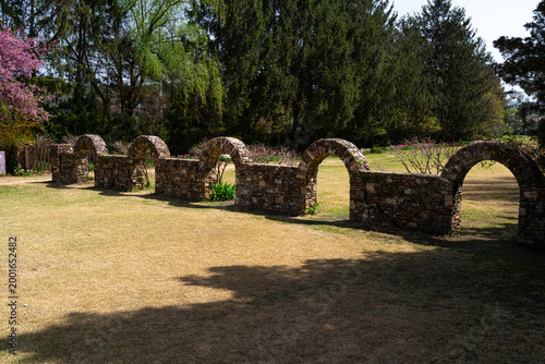 A series of rustic stone arches curves through a sunlit garden, casting deep shadows across the lawn beneath towering evergreens and vibrant pink spring blossoms.