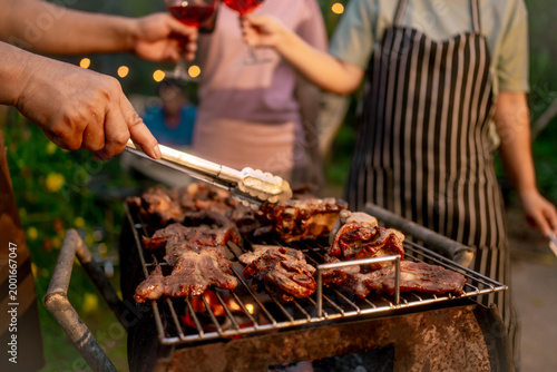 Hands grilling steaks on barbecue with tongs at outdoor garden party; people toast red wine in background, warm evening lights, casual summer gathering.