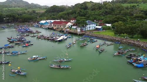 Aerial view of a traditional fishing village harbor. Many wooden boats moored in calm green water near a rural coastal settlement surrounded by lush green hills.