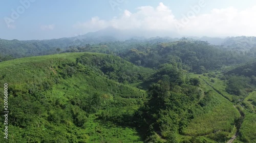 Aerial view of lush green hills and valleys with a distant mountain peak under a blue cloudy sky. Beautiful tropical nature landscape and serene environment.