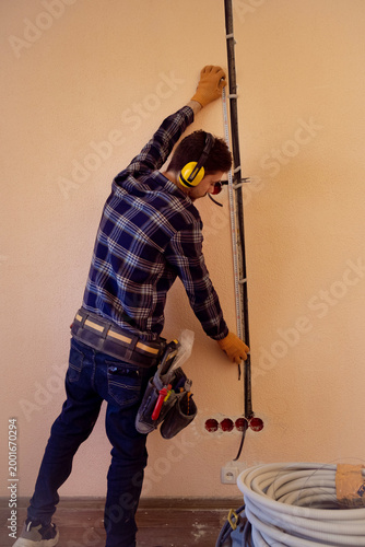 Man wearing ear protection takes measurements for precise electrical system installation in indoor construction work site