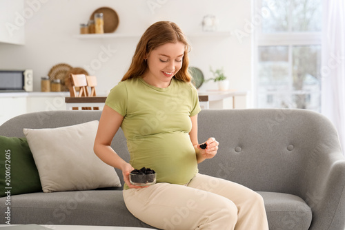 Young pregnant woman with dried prunes sitting on sofa in kitchen