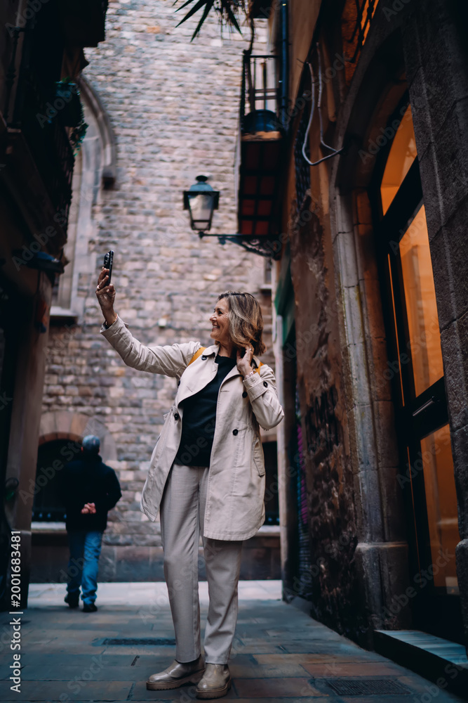 Obraz premium Full length smiling woman taking photo with mobile phone in city alley. Mature female traveler exploring urban landmarks, digital nomad lifestyle, authentic representation, copy space