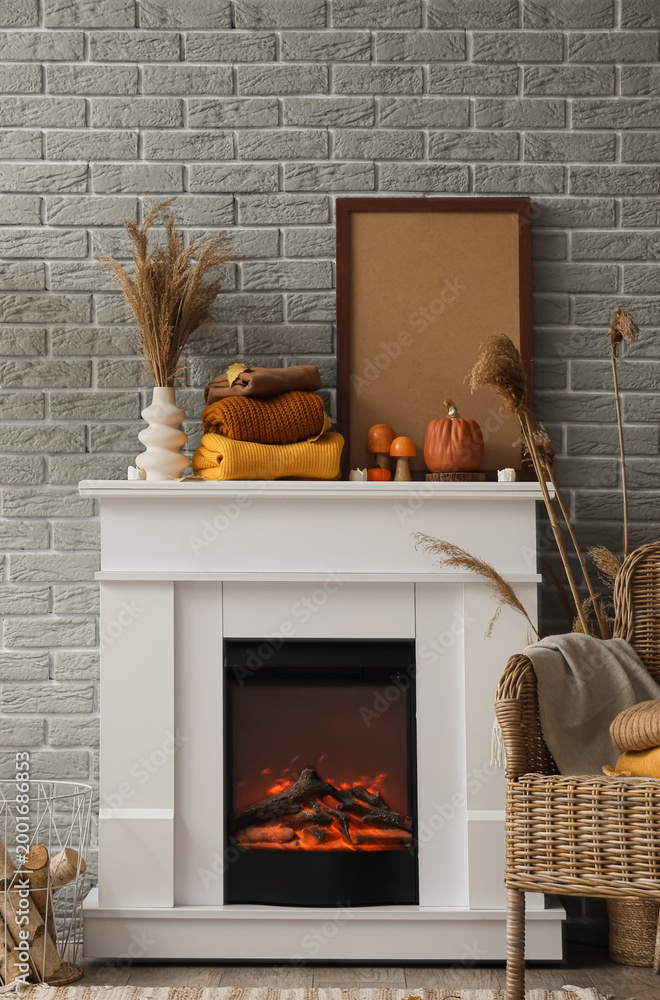 Fototapeta premium Stack of stylish warm clothes, pampas grass, autumn decor and blank picture frame on fireplace near grey brick wall