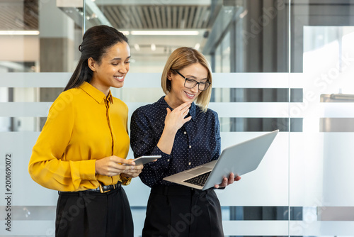 Diverse businesswomen standing together. Smiling and discussing project data on a laptop and smartphone. Showcasing teamwork and modern corporate communication within a professional office environment
