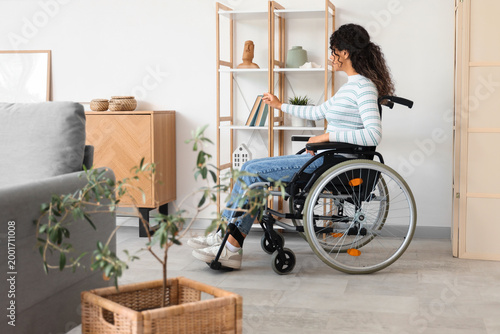 Young African-American woman in wheelchair taking book from shelf at home