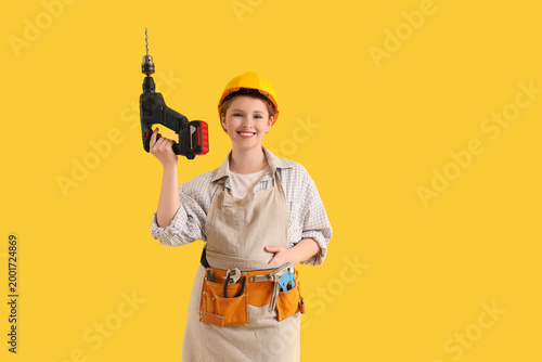 Young female carpenter with drill on yellow background. Labor Day celebration