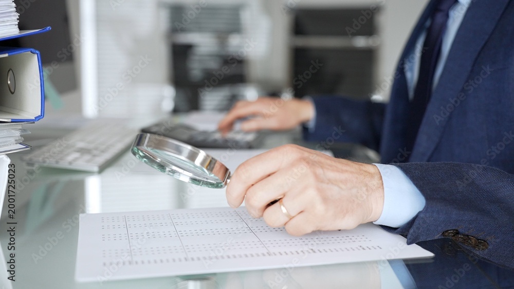 Fototapeta premium Hand holding magnifying glass, inspecting financial document, performing audit and business analysis. Audit and taxes in business