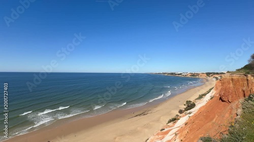 Wide panoramic view of a sandy beach with red coastal cliffs and calm sea under a partly cloudy sky.