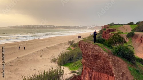 Woman smiles as she takes a selfie at the beach, enjoying the sunny weather and picturesque sea view with cliffs in the background.