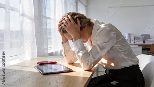 Businesswoman stressed holding head with hands in frustration while sitting at desk looking at tablet with charts worried about work problems or financial losses tired employee