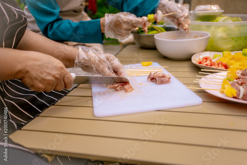 Hands preparing skewers at outdoor table, slicing yellow bell pepper on cutting board with bowls of vegetables and wooden sticks for grilling.