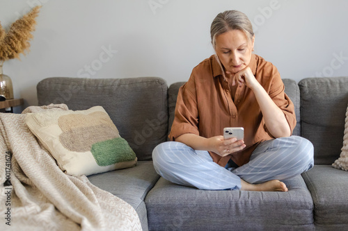 Картината върху платно Thoughtful middle aged woman using smartphone while sitting on sofa, making a decision and comparing options online