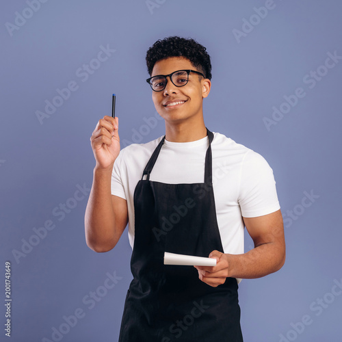 Fotografía Young African American male barista wearing black apron holds a pen and notepad
