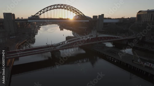 Cinematic sunrise over Newcastle showing the sun rising behind the Tyne Bridge, with the Swing Bridge and Millennium Bridge reflected in the river.