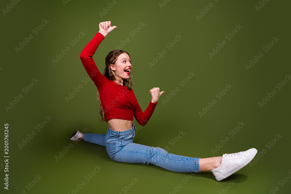 Fototapeta premium Young stylish woman in red crop top and jeans performs a joyful split against a green studio backdrop
