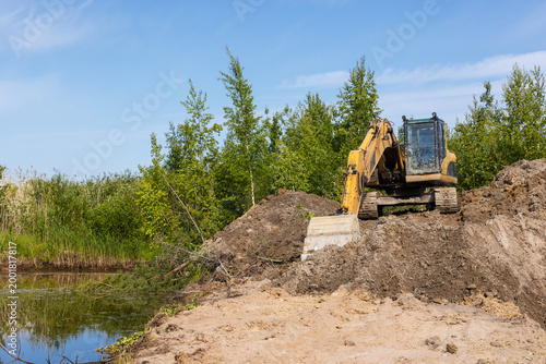 A yellow tracked excavator works along a wetland pond bank, moving soil and clearing vegetation for land development and erosion control. Bright blue sky and green trees frame the industrial scene