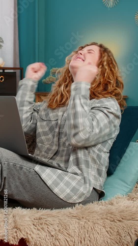 Young woman at home sitting with laptop raising hands up celebrating successful project or exciting win. Caucasian redhead girl on sofa smiling with joy, emotional reaction to achievement and success