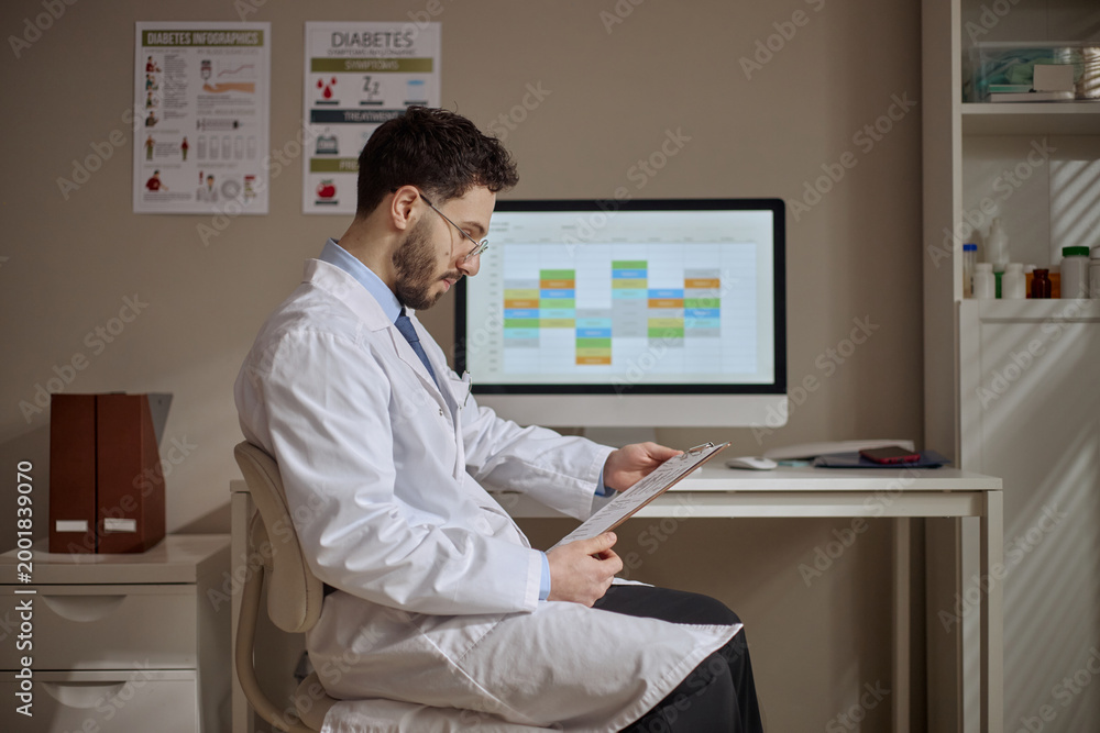 Fototapeta premium Young adult Middle Eastern male doctor reviewing clipboard at computer with schedule on screen in clinic, managing patient records. Useful for healthcare administration, medical workflow, telehealth