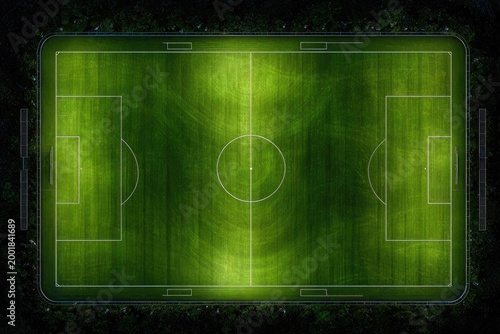 An aerial view showcases a brightly lit soccer field at night, surrounded by dense foliage, creating a striking contrast between the vibrant green and the dark environment.