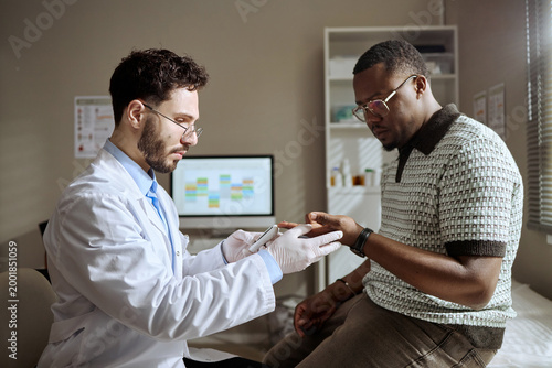 Фотография Young adult Black man receiving finger prick testing for blood glucose monitoring from Caucasian male doctor during clinic visit