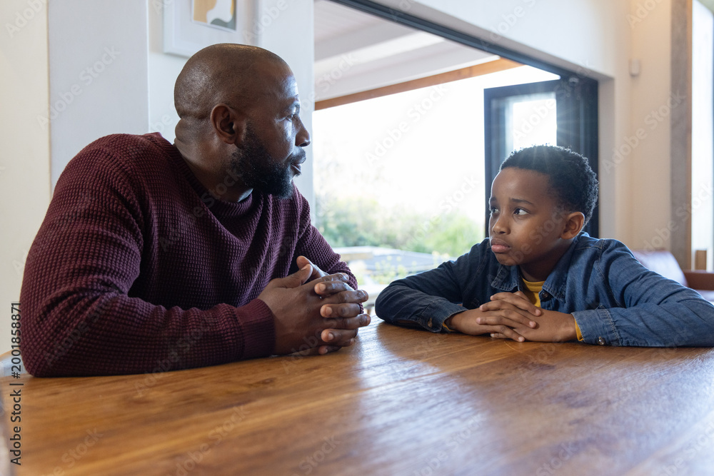 Obraz premium African American father and son sitting at wooden table near sliding door, maroon knit having talk